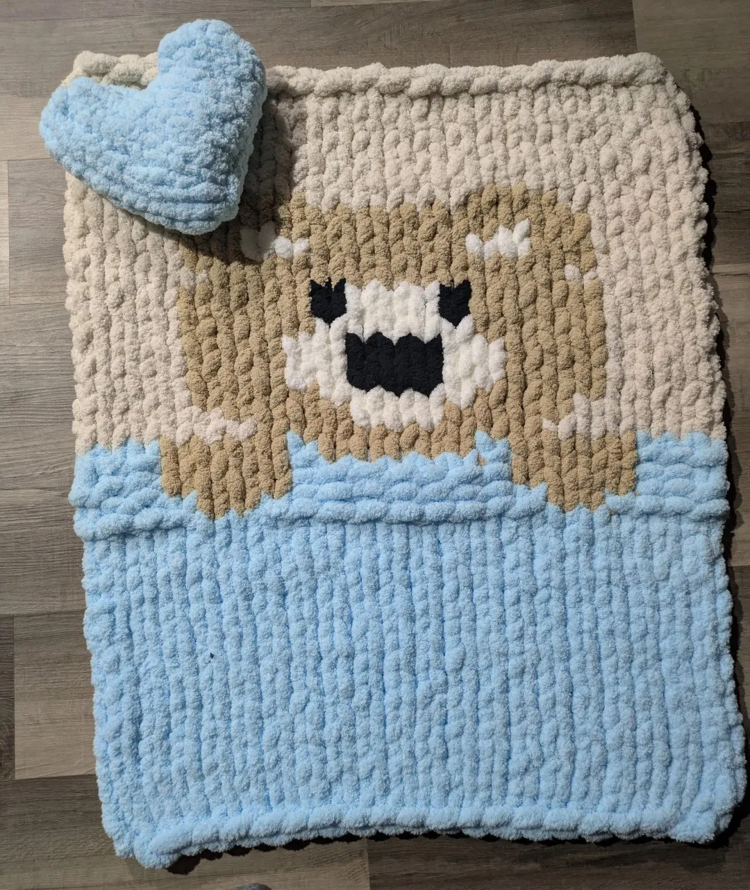 A small finger-knit blanket. It has the design of a bear tucked into a bed. In the upper-right corner is a light blue heart-shaped pillow, also finger-knit