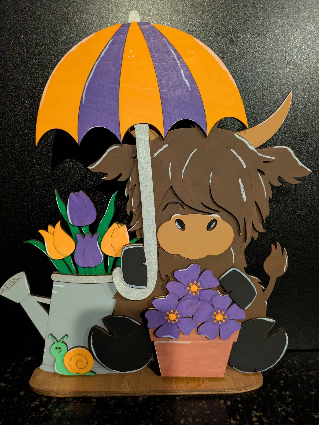 A flat wooden figure depicting a furry cow holding an umbrella. There is also a flower pot, a watering can with tulips, and a snail.